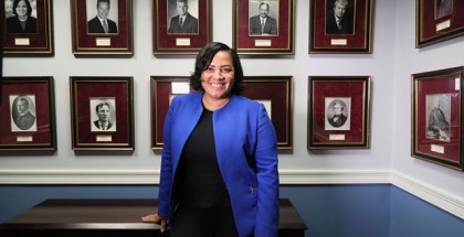 A woman in a blue suit jacket and black pants and shirt smiles at the camera while leaning on a desk in front of a wall of portraits