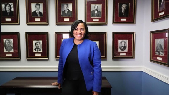 A woman in a blue suit jacket and black pants and shirt smiles at the camera while leaning on a desk in front of a wall of portraits