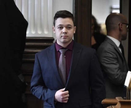 A young man in a black suit jacket and red shirt walking forward in a courtroom
