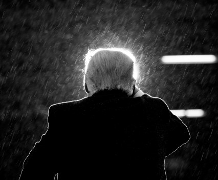 A shot of Donald Trump from behind, his head lit by a light while rain falls