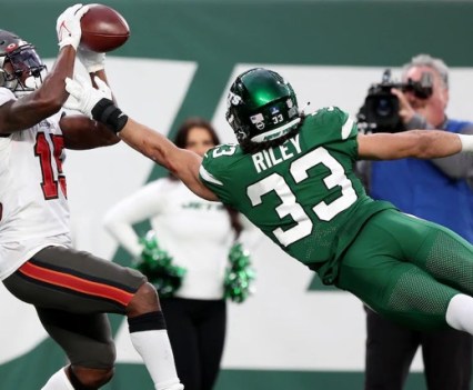 A football player in all green is mid-jump, reaching for the football that's landing in another player's hands