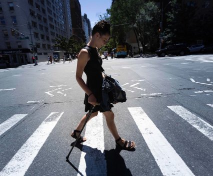 Rebecca crosses a New York City street on a hot June day, wearing sleeveless dress and sandals, using her cane. She looks down at the crosswalk.