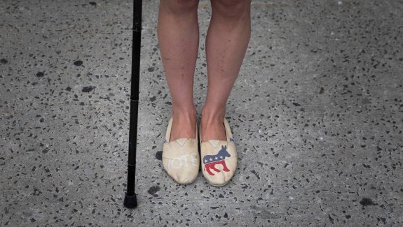 A photo of Rebecca from the knees down. She stands with her cane, wearing slip-on shoes with a democratic donkey on the left shoe and Vote written on the right shoe.