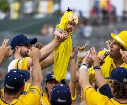 A baseball team in all yellow is cheering as a baby in a banana costume is raised above their heads