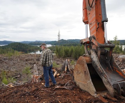 A man stands next to a crane, looking out at a large area of clear cut forest