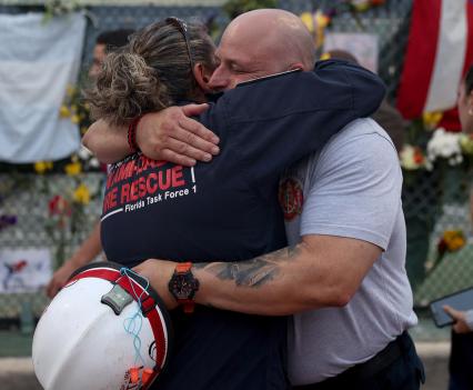 Two firefighters hugging in front of a memorial on a fence outside Surfside condo