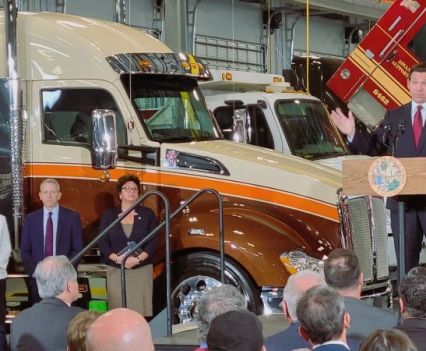 A group of people gathered near large trucks to listen to a man speaking from a high podium