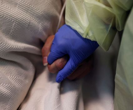 A woman in her personal protective equipment (PPE) gear holds the hand of a COVID-positive patient at a hospital