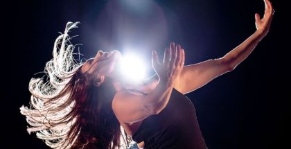 A woman with outstretched arms leans back with her hair flying out from her head. There is a bright light shining right above her.