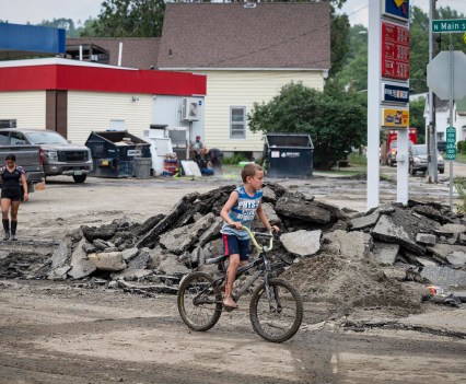 Photo of a young boy riding his bike in front of a destroyed road in a Vermont town following the flood