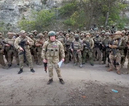 Photo of a man in front of a group of men, all in military uniforms, facing the camera as man holds gun in one hand and paper in the other