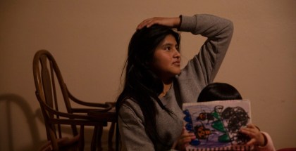 Photo of young woman with a hand on her head seated behind a child who is coloring