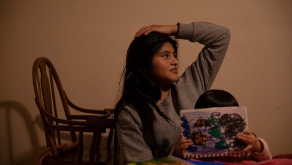 Photo of young woman with a hand on her head seated behind a child who is coloring