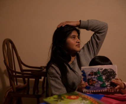 Photo of young woman with a hand on her head seated behind a child who is coloring