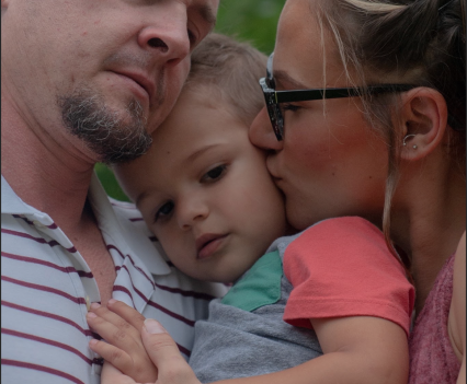 A man and a woman hold a young child between them. The man rests his head on the child's head. The woman is kissing the child's cheek.