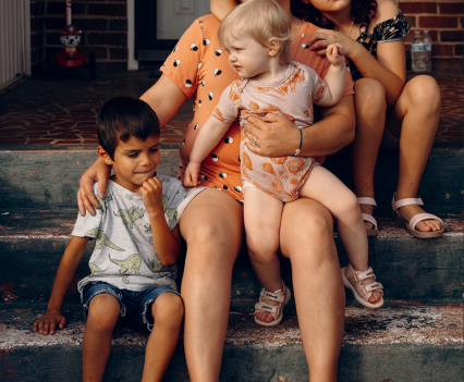 A woman sits on stairs. She has a toddler on her lap and her arm around a young boy sitting beside her. There is a young girl sitting behind the woman with her arms around the woman's shoulders.