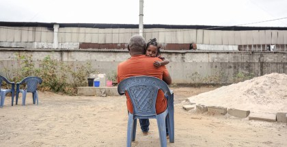 A man in an orange shirt sits in a plastic lawn chair. A young child sits in his lap and looks at the camera over his shoulder