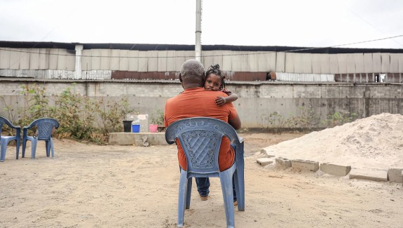 A man in an orange shirt sits in a plastic lawn chair. A young child sits in his lap and looks at the camera over his shoulder