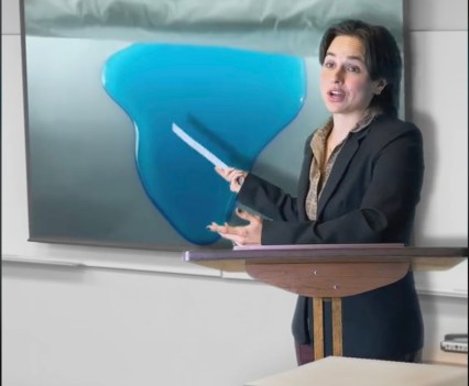 A woman in a business suit stands in front of a screen pointing to what appears to be an image of blue liquid on the screen