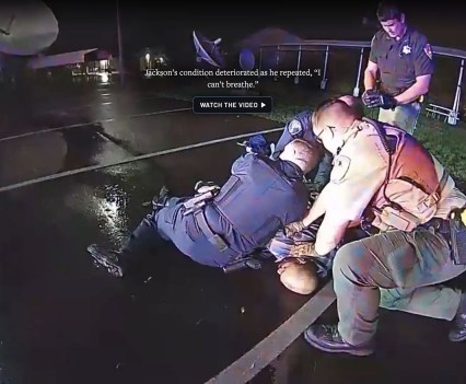 Three uniformed officers restrain a man in the prone position on the ground while another officer stands nearby