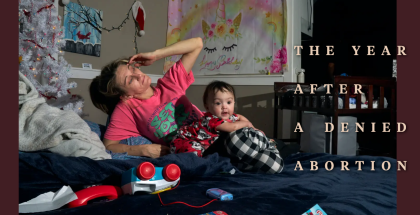 A woman in a pink shirt reclines on a bed with toddler in her lap