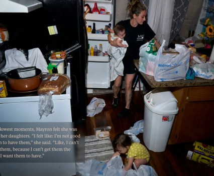 A woman holding a baby is taking groceries out of bags and putting them into the refrigerator while a toddler plays on the floor