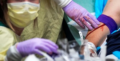 A woman in a yellow mask and medical cape wears bloody purple gloves while administering a blood transfusion to a patient