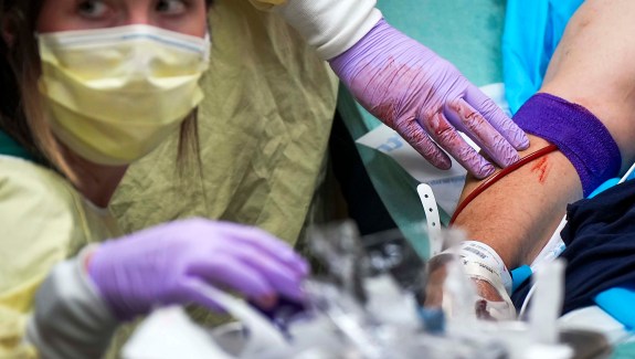 A woman in a yellow mask and medical cape wears bloody purple gloves while administering a blood transfusion to a patient