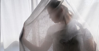 A teenager wearing a teeshirt poses in front of a sunny window. She is in front of a sunny window, under a sheer curtain. Her right hand is out is front of her as if she is pushing through the material, and her face turned to look at her hand. Only a profile of her face is visible under the curtain.