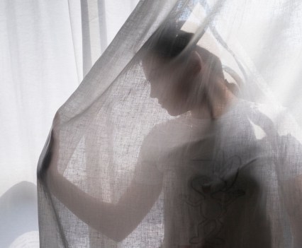 A teenager wearing a teeshirt poses in front of a sunny window. She is in front of a sunny window, under a sheer curtain. Her right hand is out is front of her as if she is pushing through the material, and her face turned to look at her hand. Only a profile of her face is visible under the curtain.