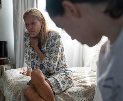 A teenager in a teeshirt is in the foreground, angled slightly away from the viewer and blurry. He mother is sitting on bed behind her. She is in a floral dress, legs crossed, and right arm resting across her chest to her neck. She is crying and leaning to the left.