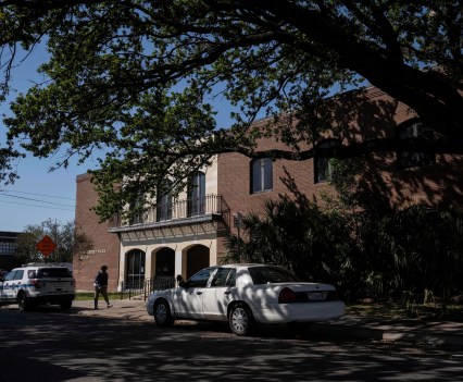 A brick police station is partially shaded by the branches of a large tree on the opposite side of the street from the viewer. Three police cars are parked by the curb in front of the building.