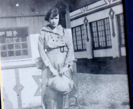 A black and white photo of a woman standing alone outside. She pinches the floppy brim of her hat between her fingers and stares at the camera.