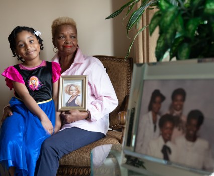 Photo of an elderly Black woman with a younger Black girl on her lap. The elderly woman holds a framed photograph of an individual, and in the forefront is another photo of a family