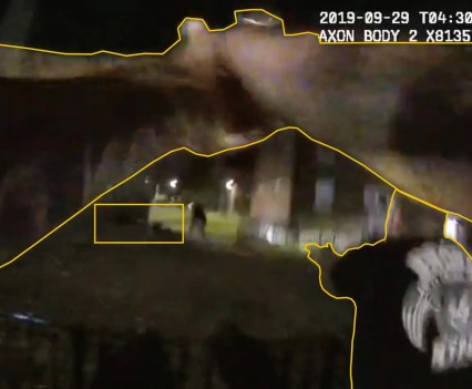 Still image from blurry body cam has yellow lines drawn on top of the blurry image to trace what appears to be hands holding a gun pointed at a person