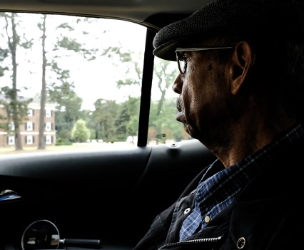 A man sits in a car looking out at buildings in the distance